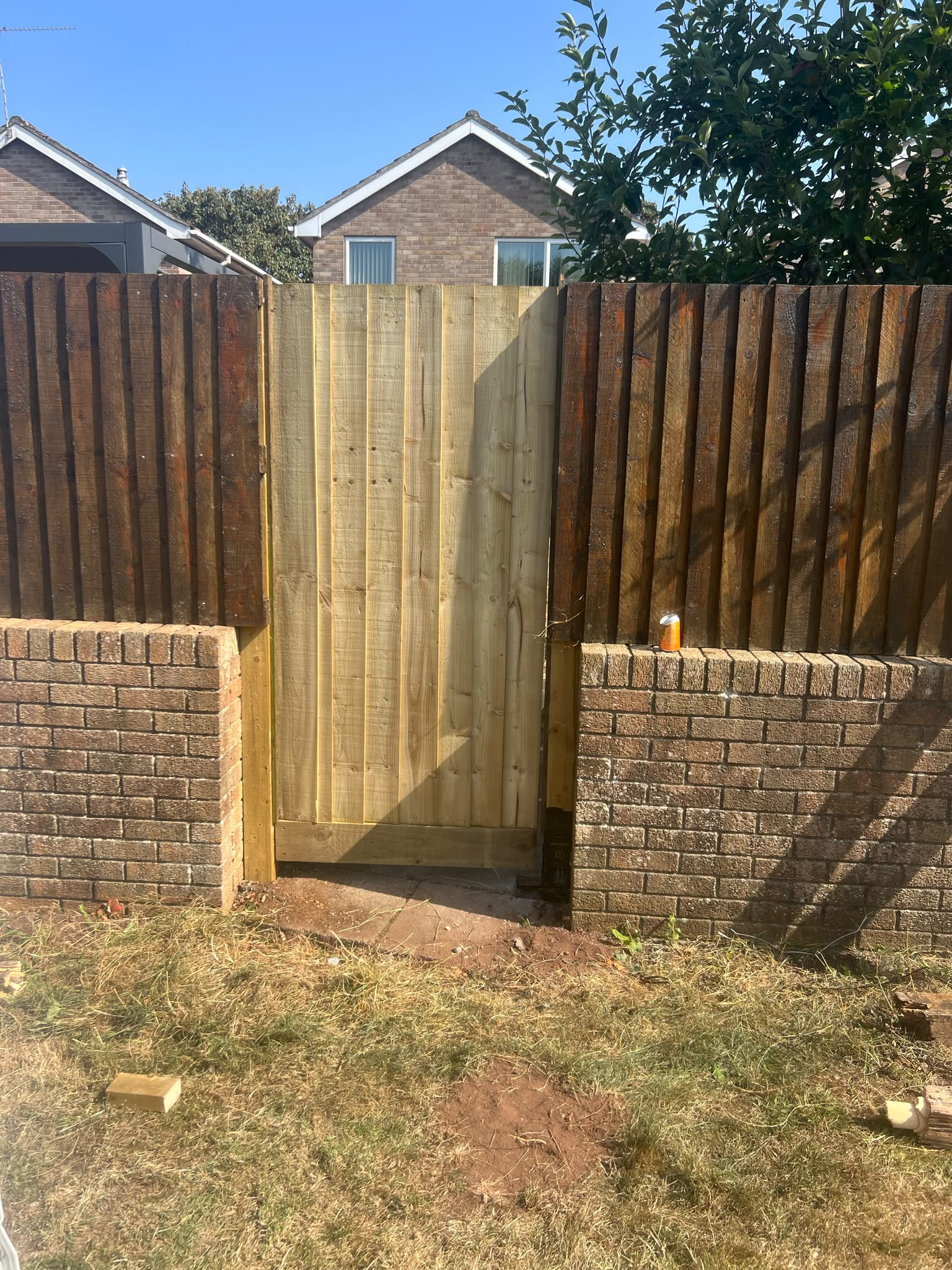Light wooden closeboard gate hung between darker fence panels on a low brick wall with houses beyond