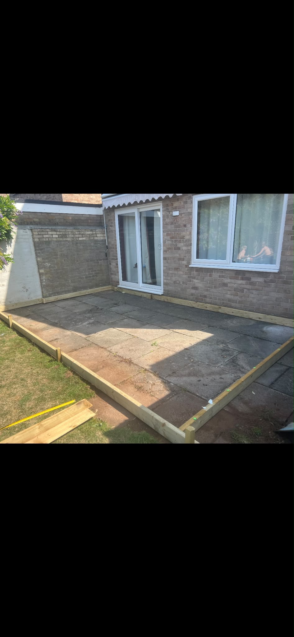 New timber frame staked around weathered paving slabs beside a brick house with a measuring tape on the lawn