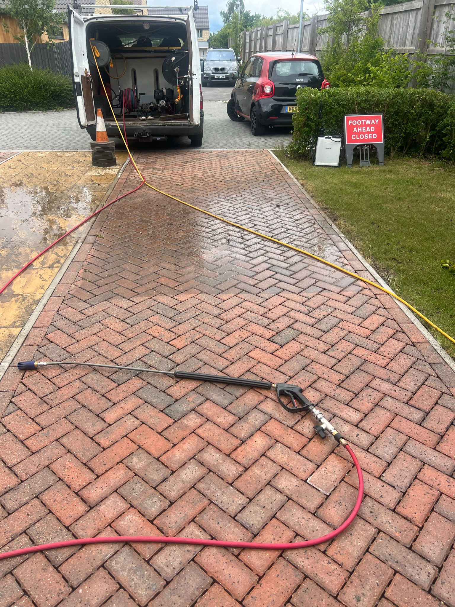 Red herringbone block driveway being cleaned with a pressure washer lance, red hose, and open white van