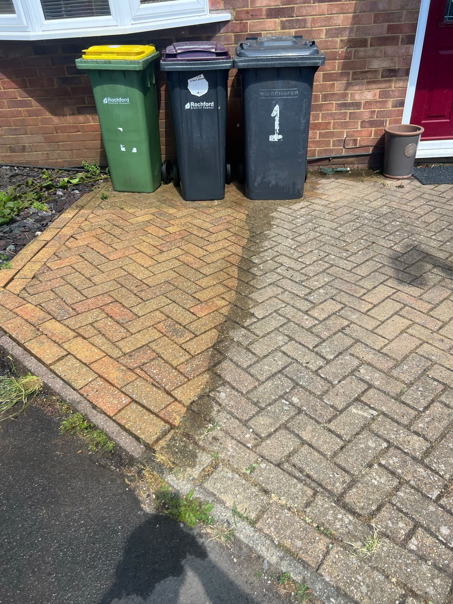 Herringbone driveway showing clean bright orange paving beside dirty grey paving with wheelie bins against a brick wall