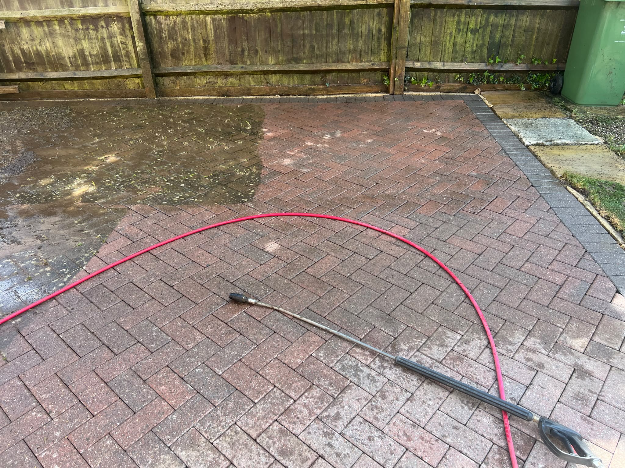 Red herringbone patio with cleaned paving, pressure washer lance and red hose, while a corner remains algae-stained