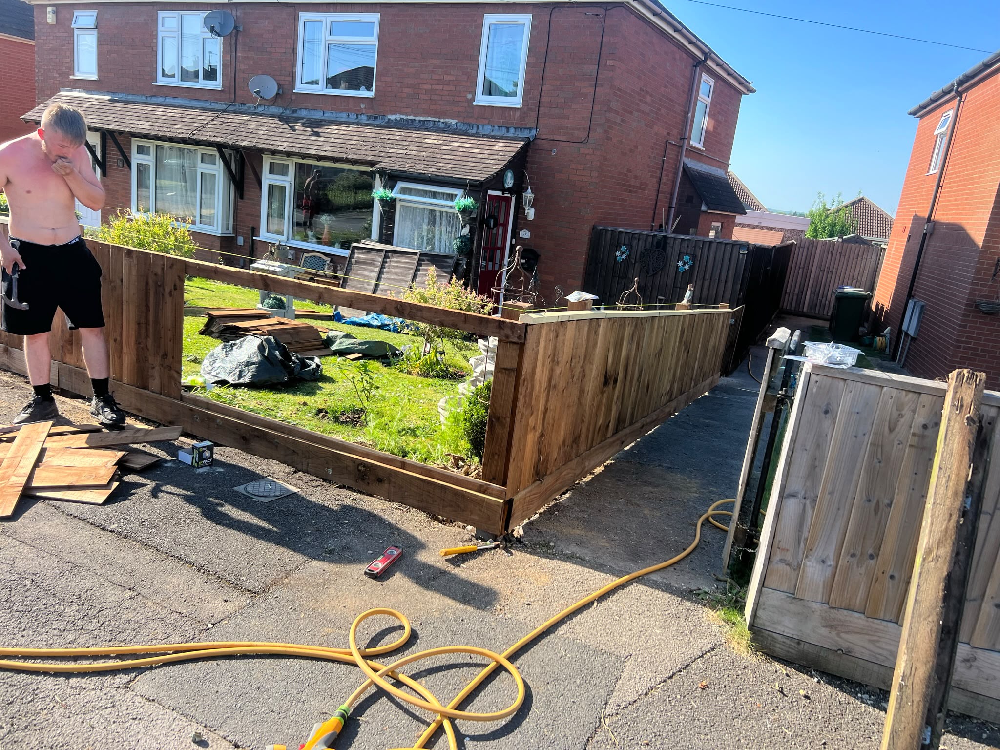Worker hammering dark vertical boards onto a timber fence frame around a front lawn on a sunny street