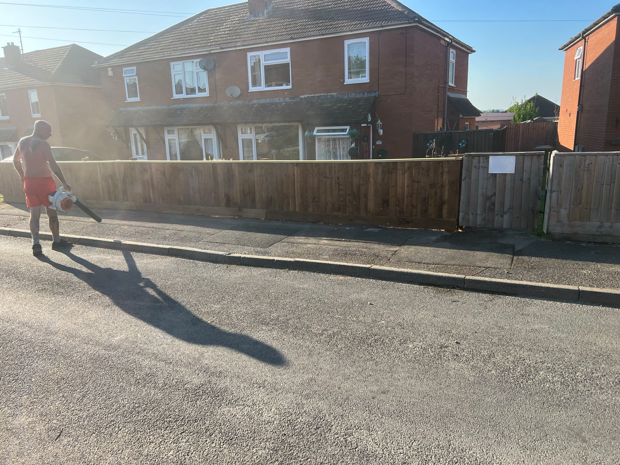 Newly installed light closeboard fence and gate with a worker leaf-blowing dust from the pavement