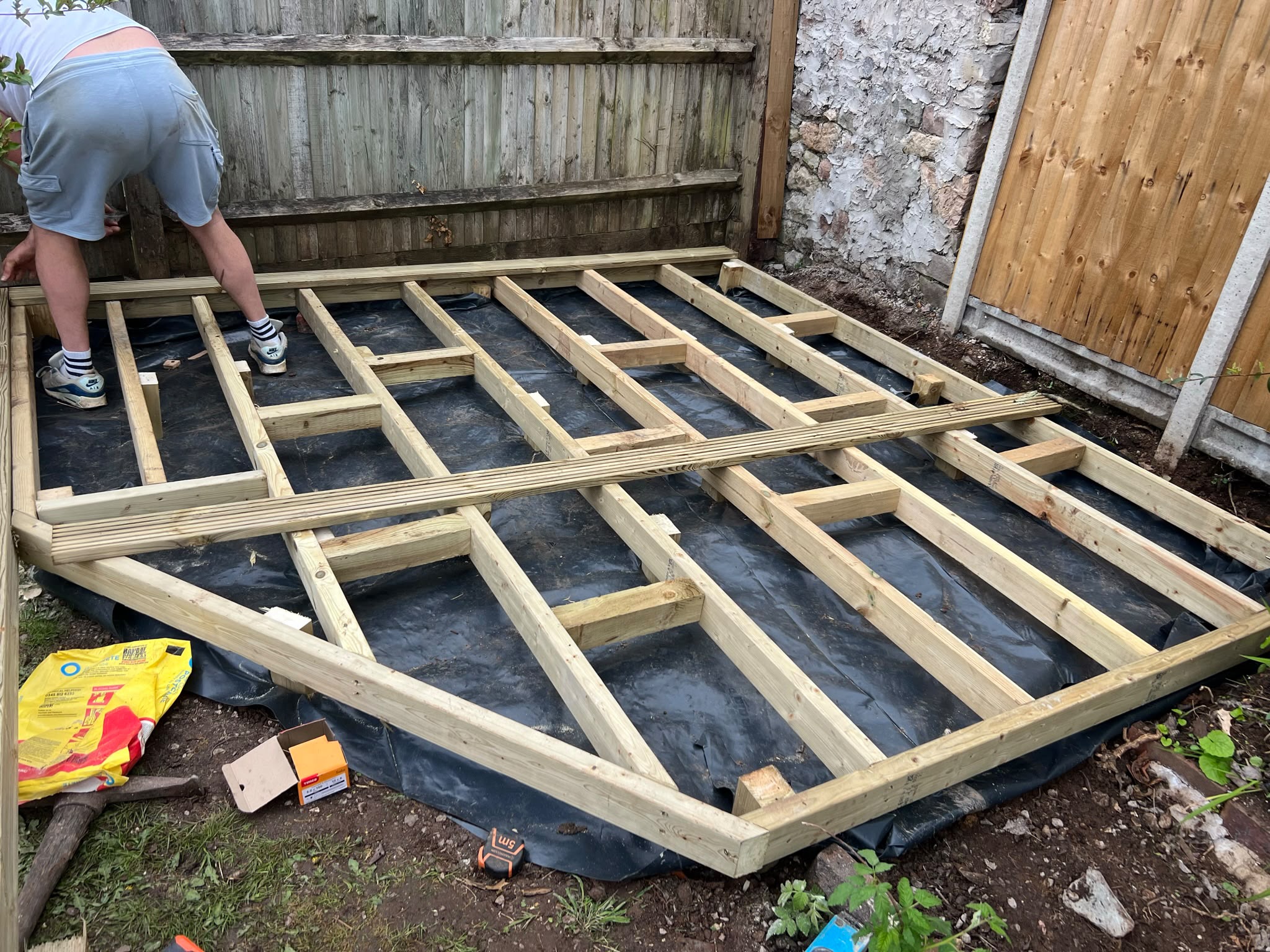 Worker assembling a timber deck frame over black weed membrane beside wooden fencing and a stone wall