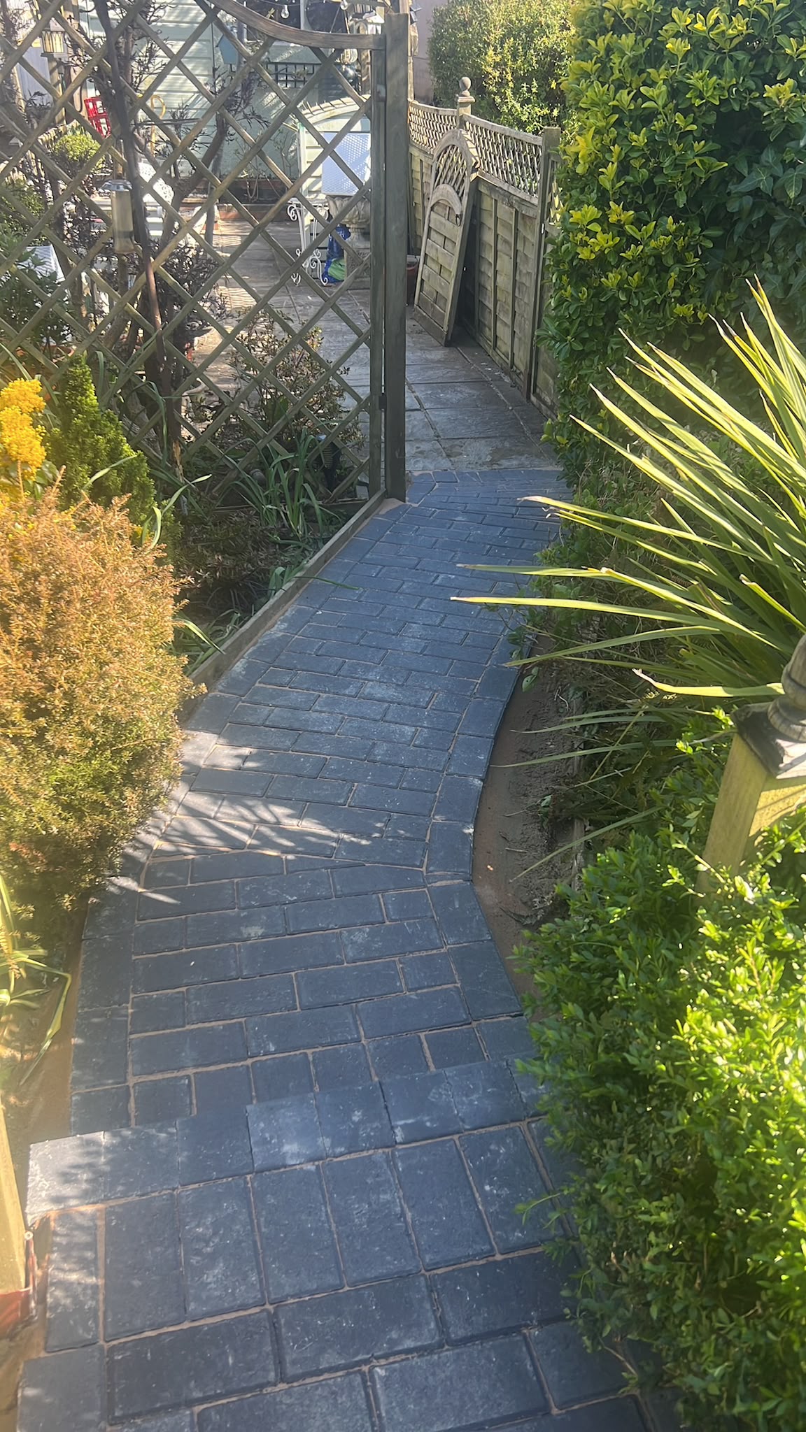 Newly laid dark charcoal block path curving through garden planting toward an open wooden gate