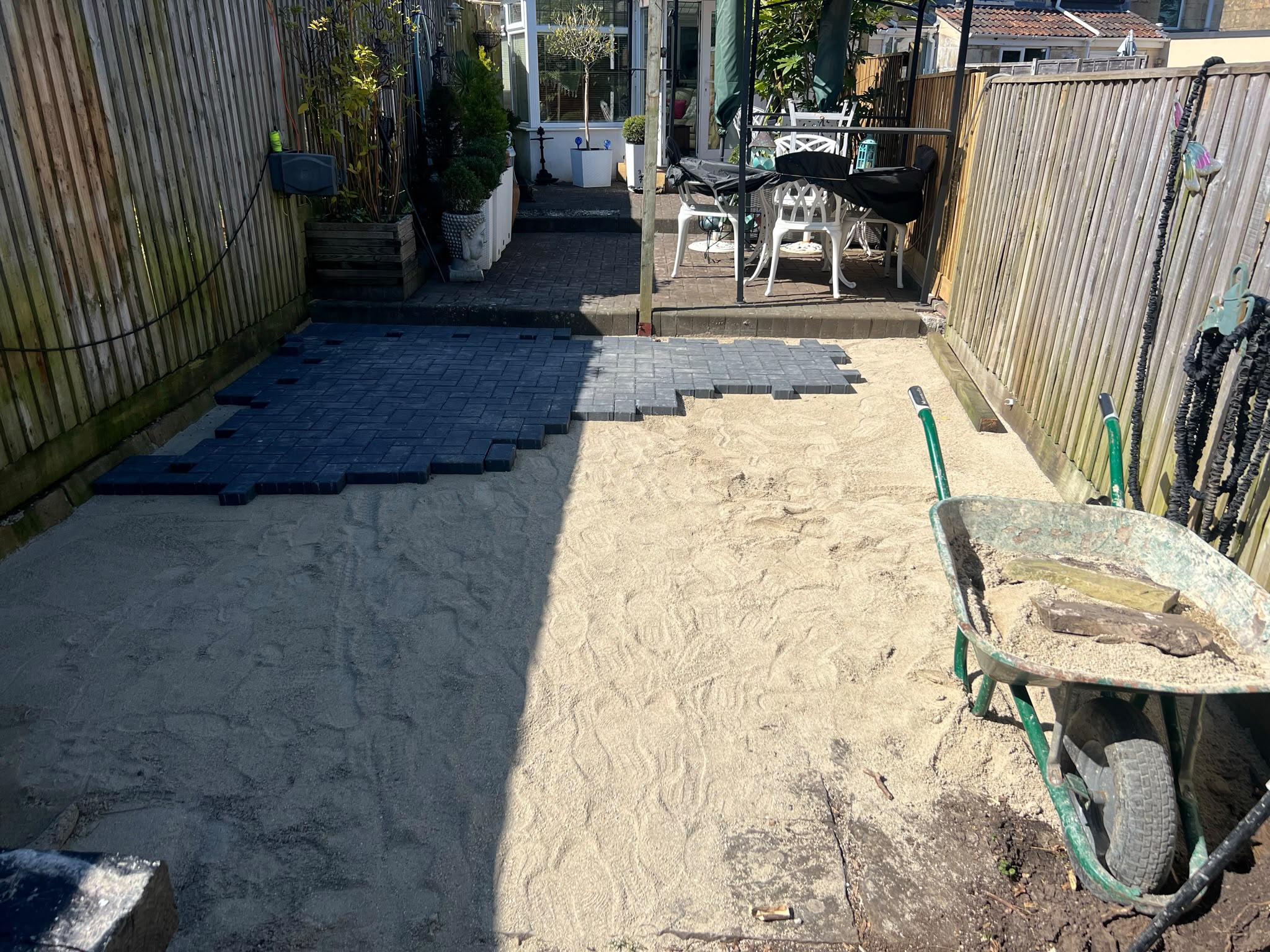 Sand base with dark charcoal block paving being laid between wooden fences, green wheelbarrow in the foreground