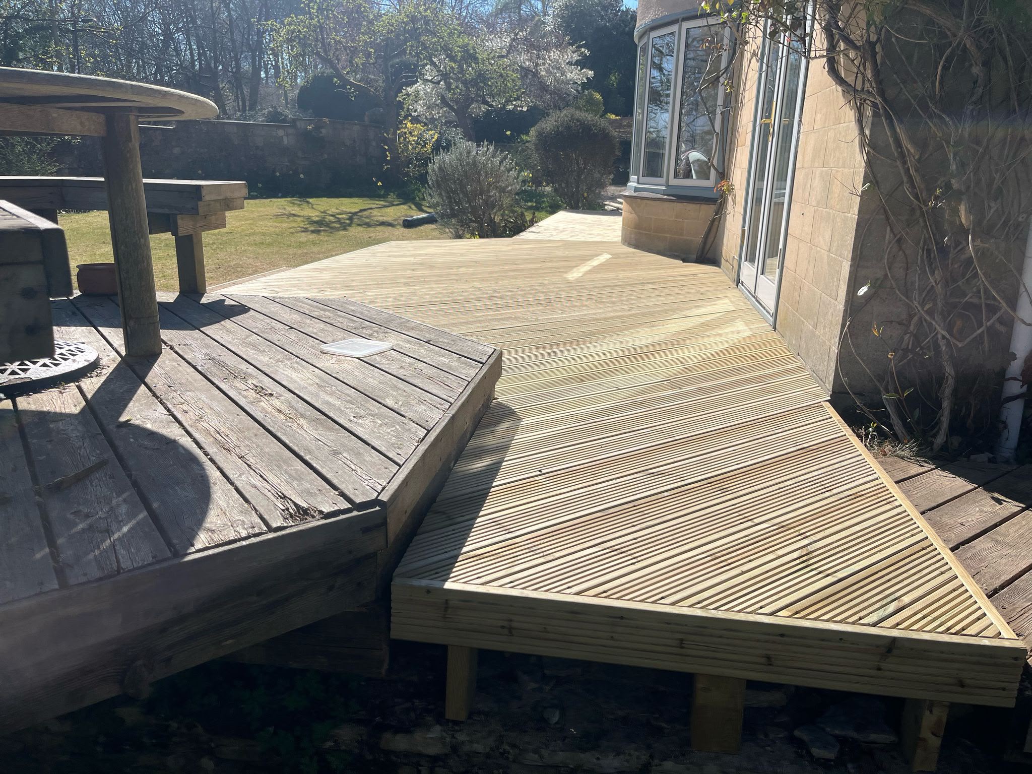 New light timber multi-level deck with diagonal boards beside a stone house and older weathered wooden seating