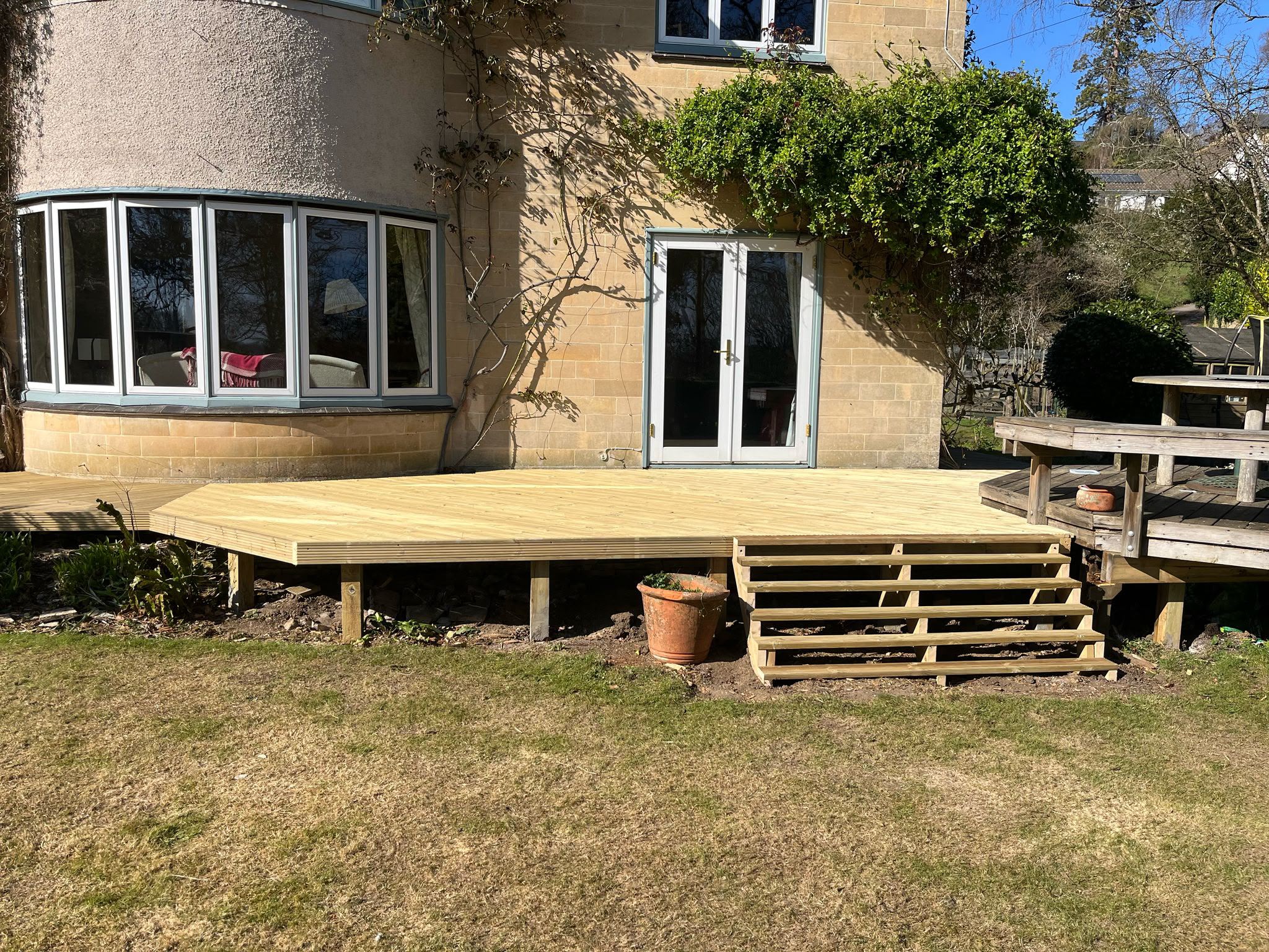 Fresh light timber deck with five open-riser steps outside a stone house with bay window and French doors