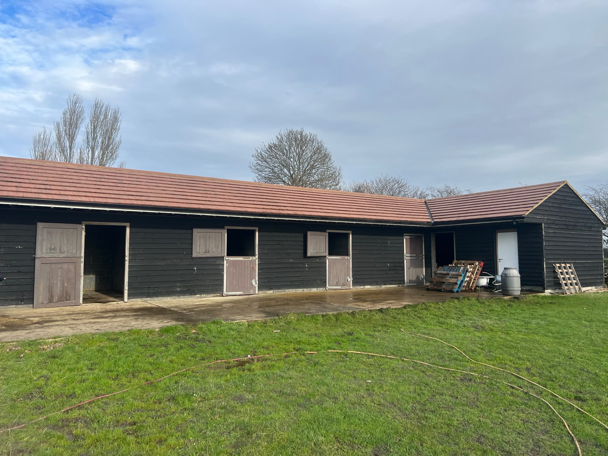 Black wooden stables with a wet cleaned concrete apron and yellow hose across the lawn