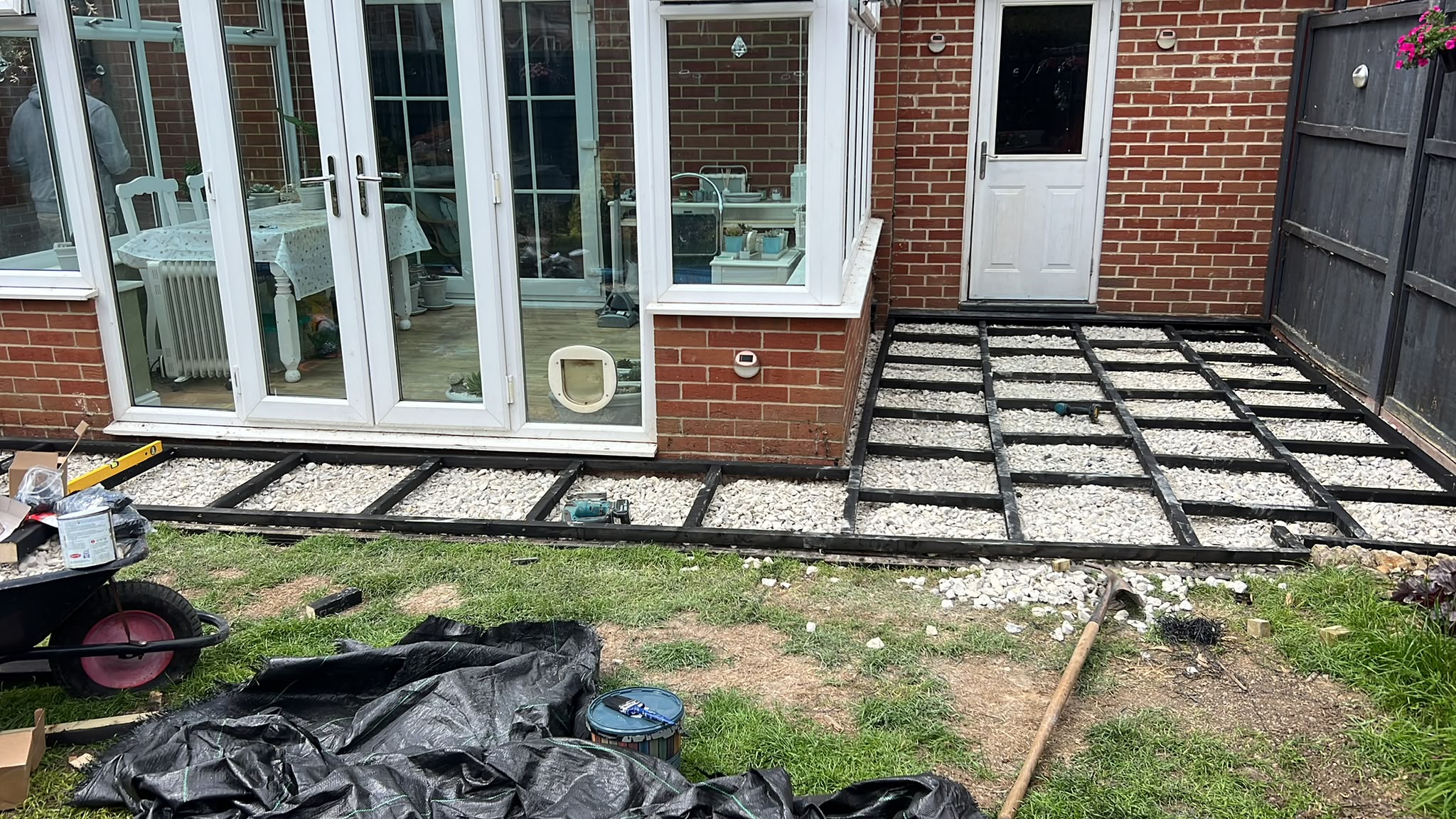 Black-painted timber deck subframe on white aggregate beside a brick house and conservatory with tools on the lawn