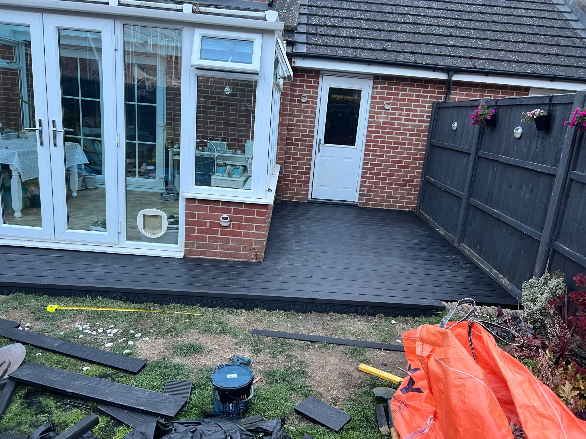 New black composite decking at a conservatory doorway with spare boards, tools, and an orange bulk bag on the lawn