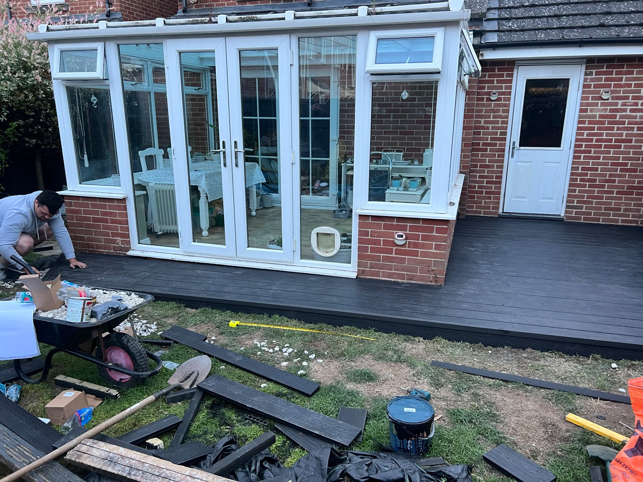 Worker fitting dark composite deck boards around a white conservatory with tools and a wheelbarrow on the lawn