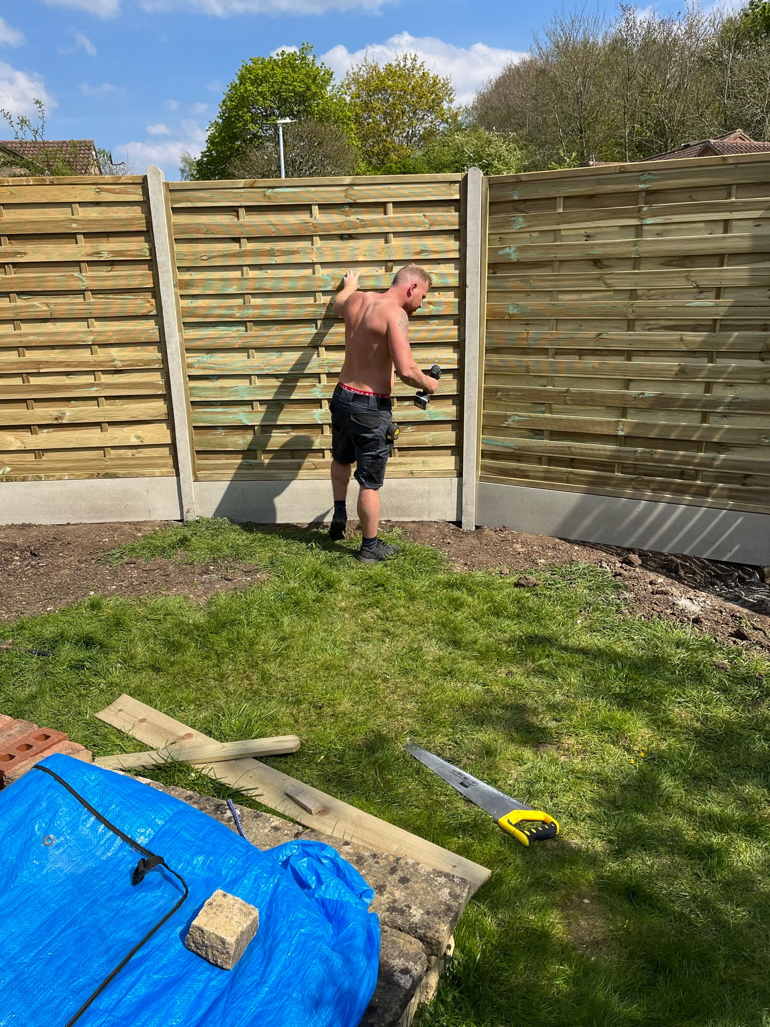 Worker with a power drill fitting horizontal wooden fence panels into concrete posts on a sunny day