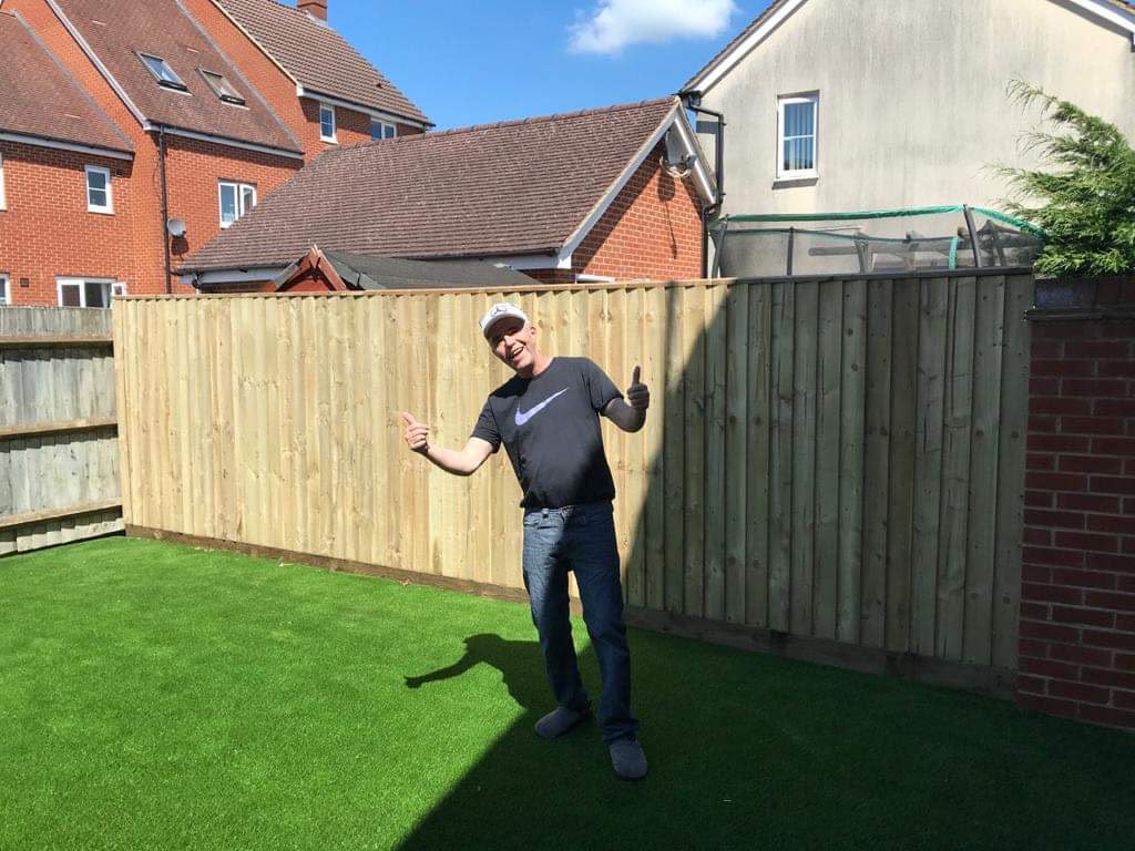 Man giving thumbs up in front of a new light wooden fence on green artificial grass under blue sky