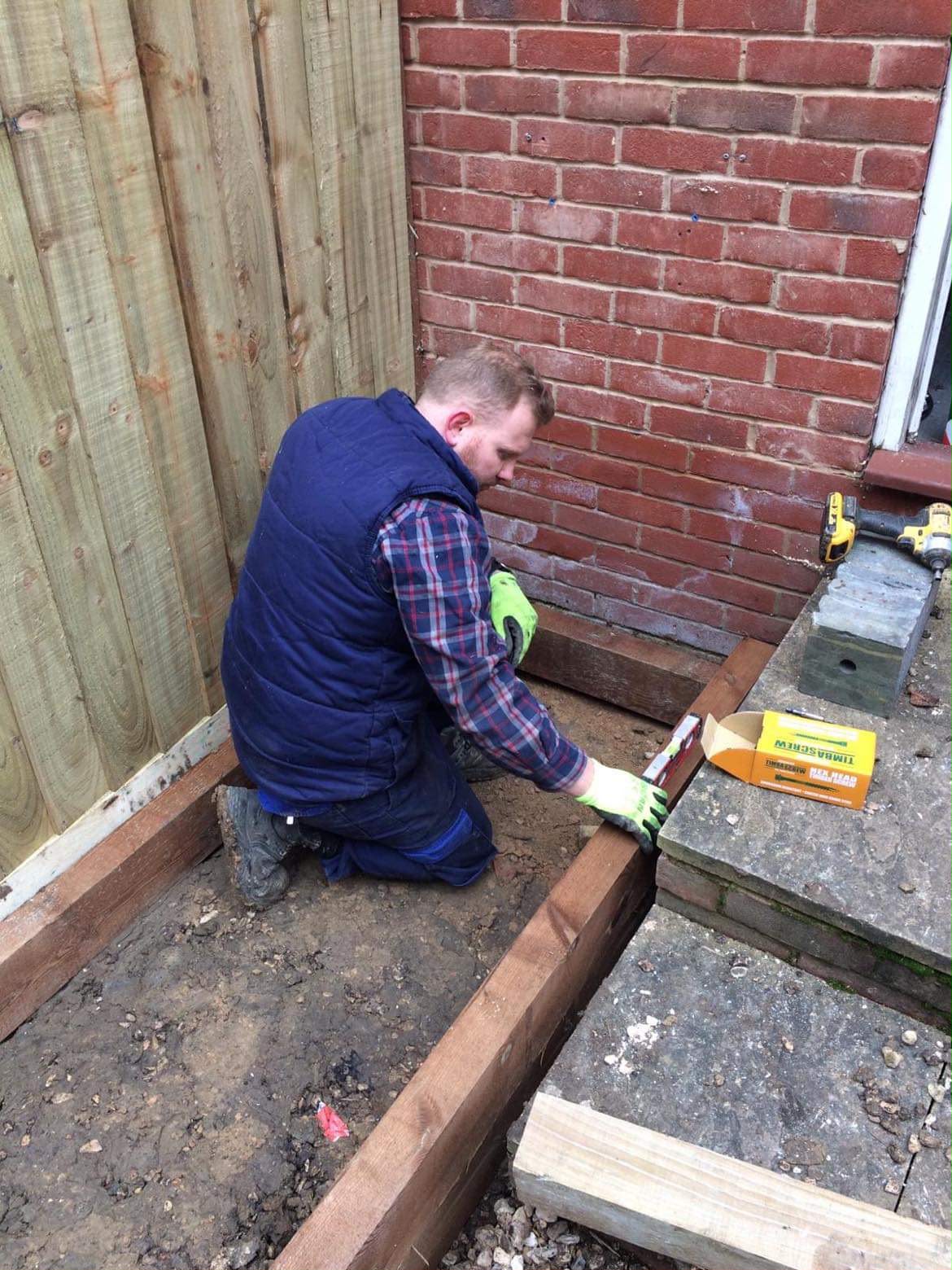 Worker using a spirit level to align timber sleepers for a retaining border beside a new fence and brick house