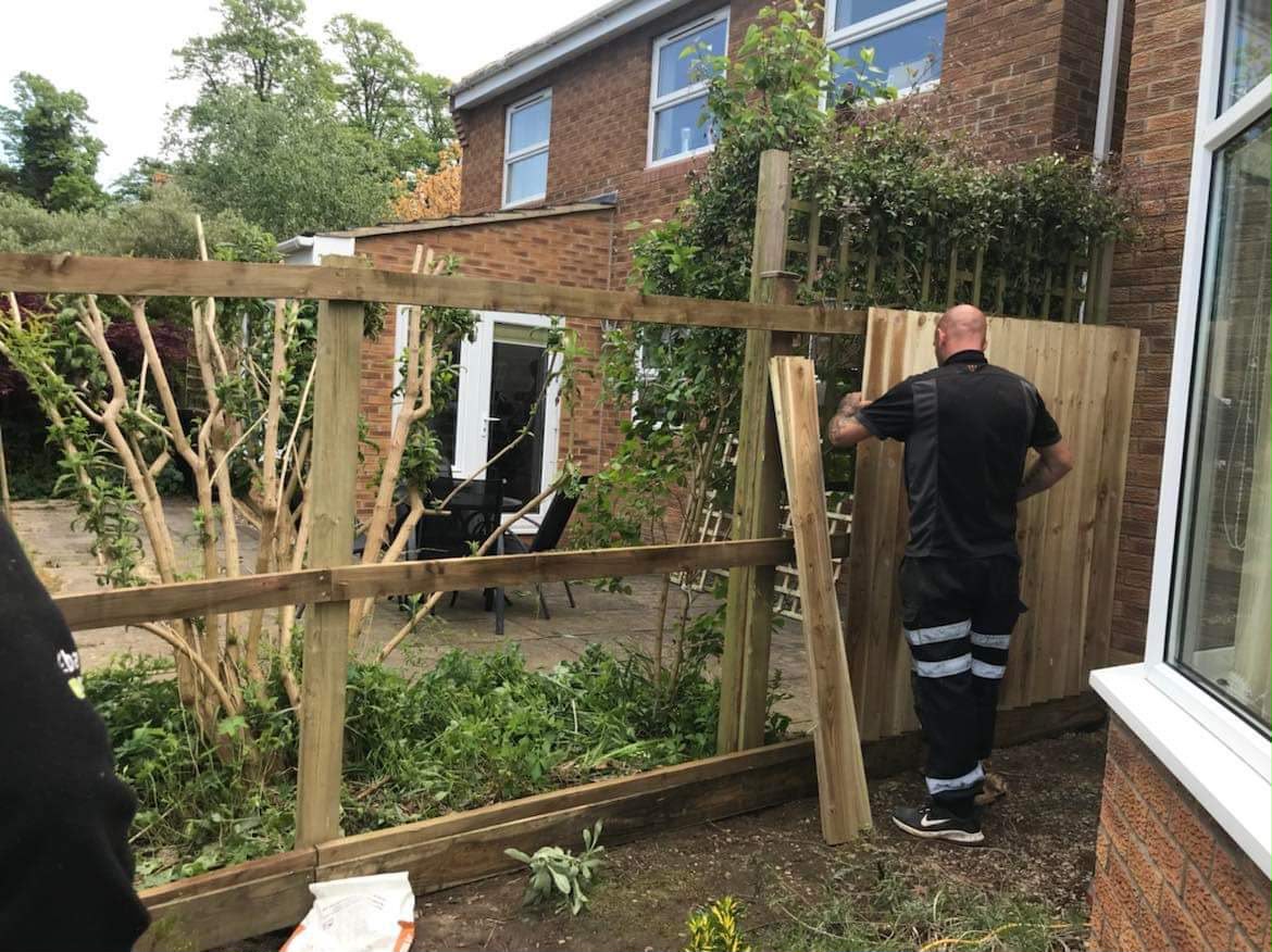 Worker attaching vertical timber boards to a fence frame of posts and rails beside a brick house