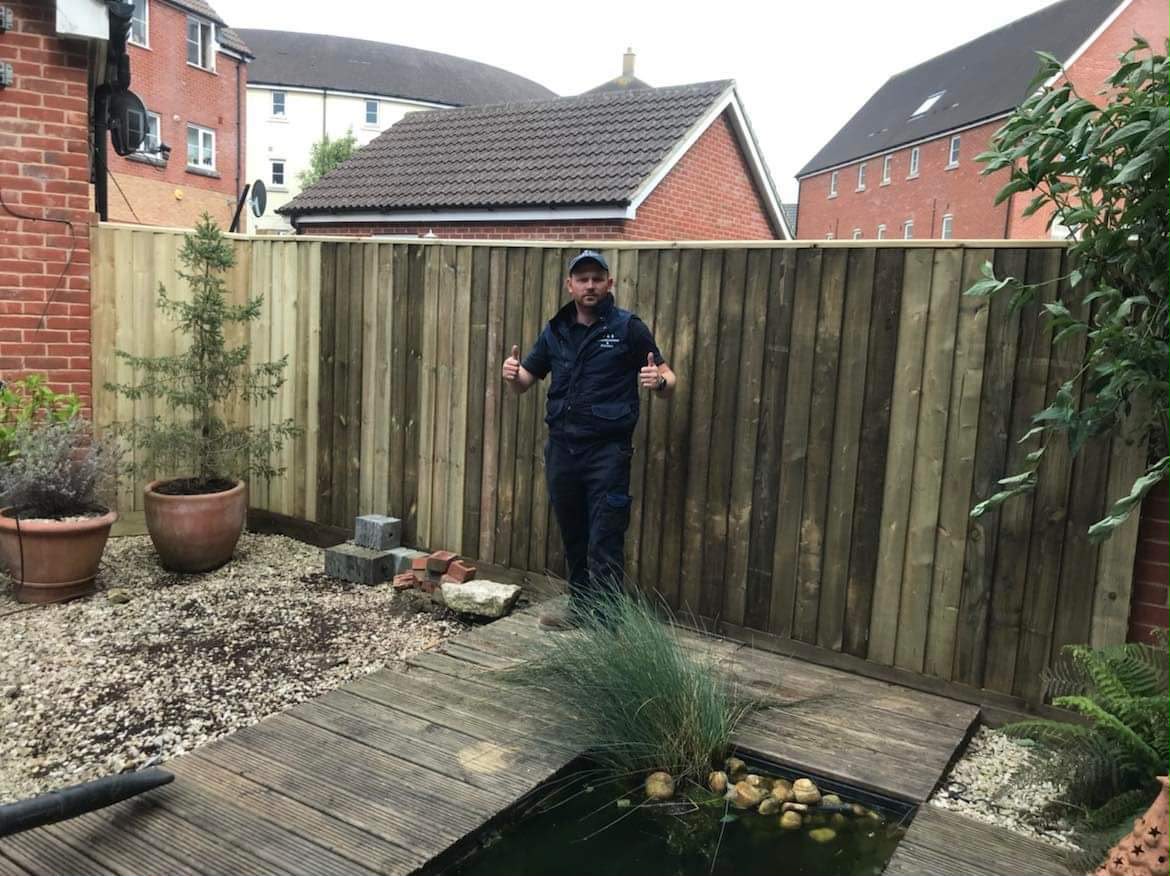 Worker giving double thumbs up in front of a new tall closeboard fence beside decking, pond, and pots