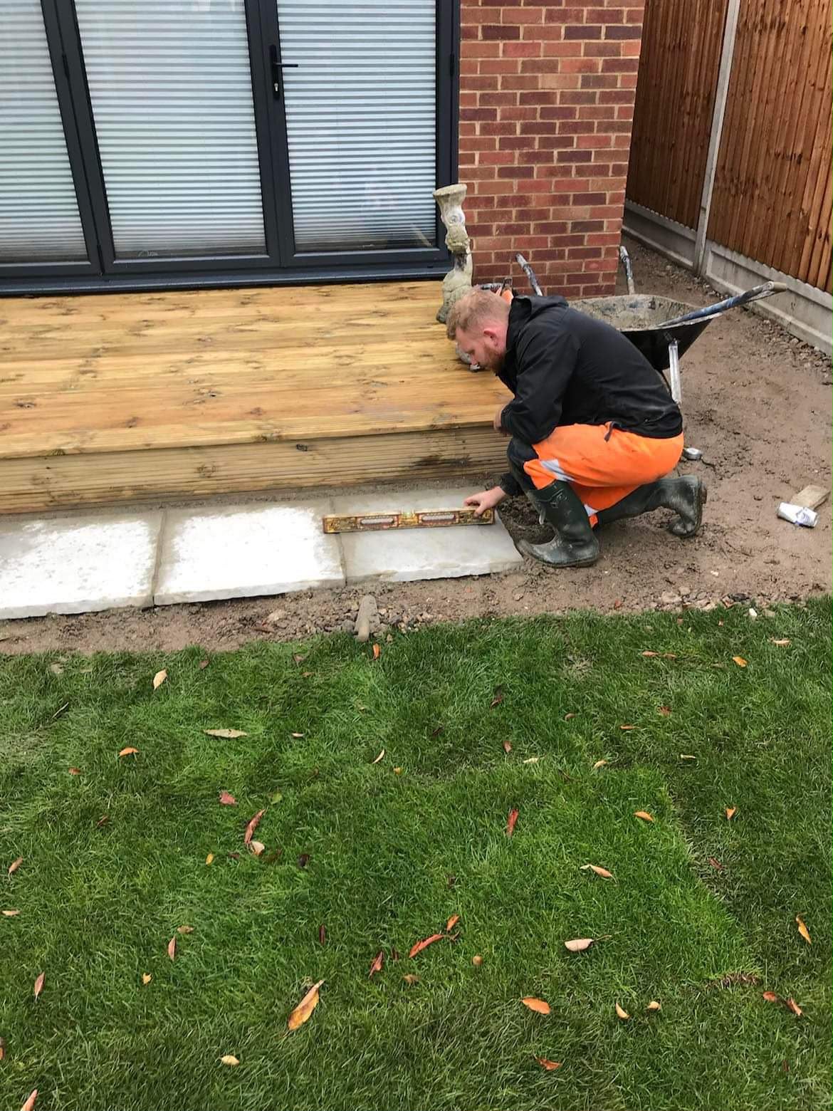 Worker kneeling with a spirit level on light grey patio slabs beside timber decking and a wheelbarrow of mortar