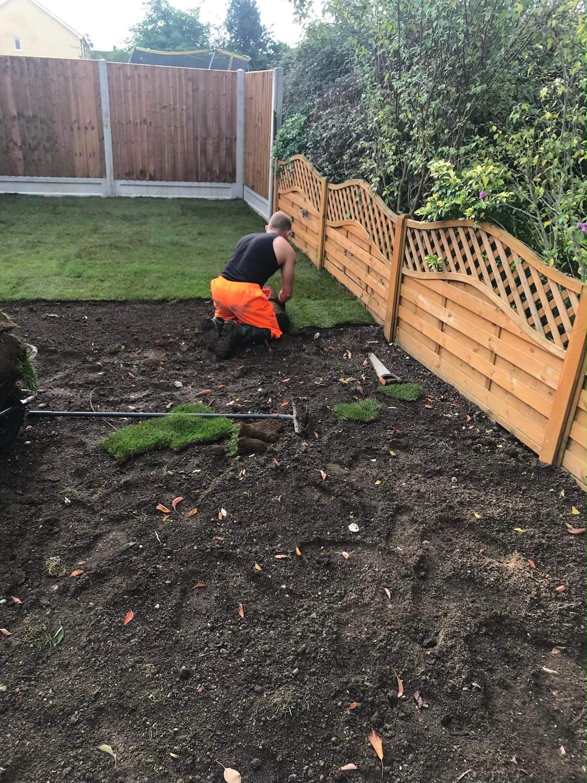 Worker in orange trousers kneeling to lay green turf rolls on prepared dark soil beside wooden fencing