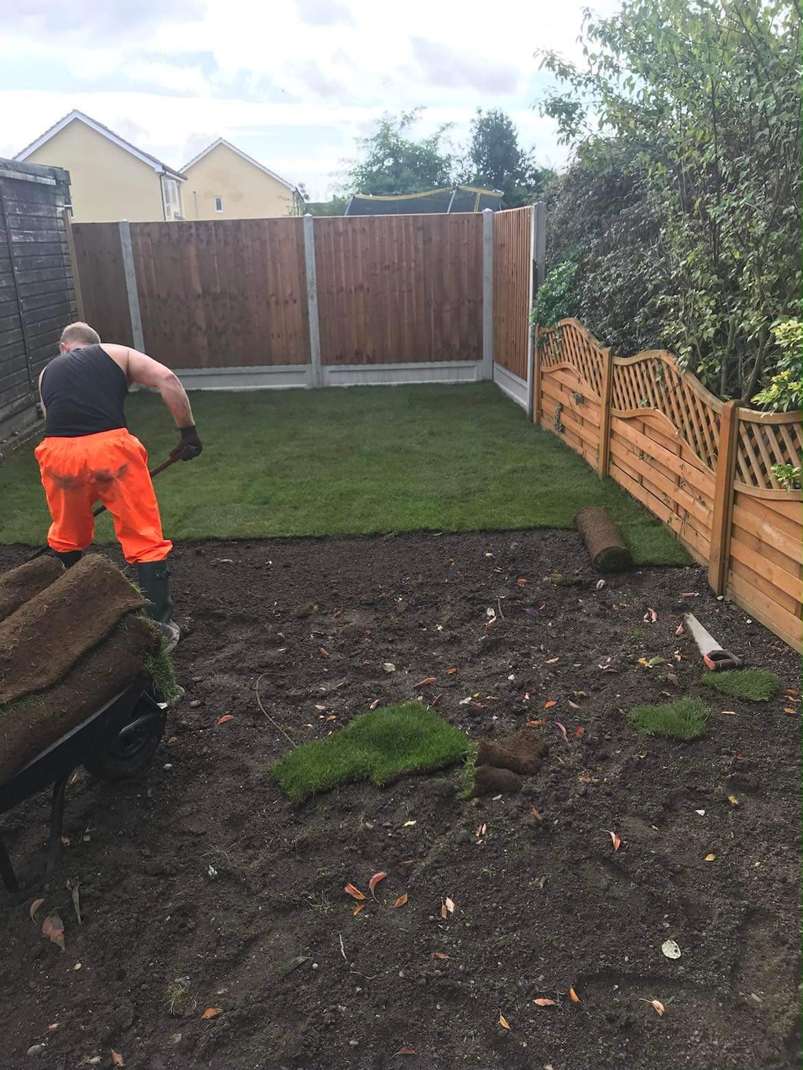 Worker laying turf with a wheelbarrow of grass rolls, half the lawn finished and half bare soil