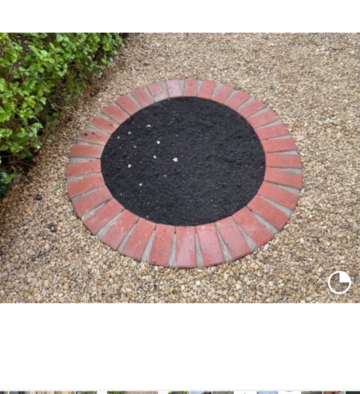 Circular planting bed edged with radial red bricks filled with dark soil set in tan gravel beside a trimmed hedge